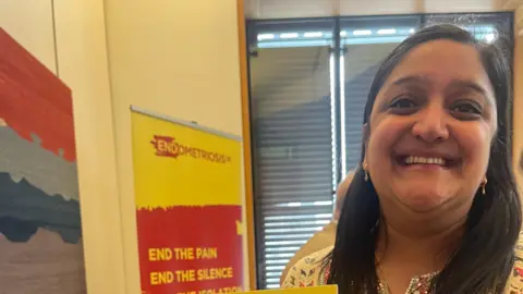 Sanju Pal smiles into the camera in front of a poster about endometriosis