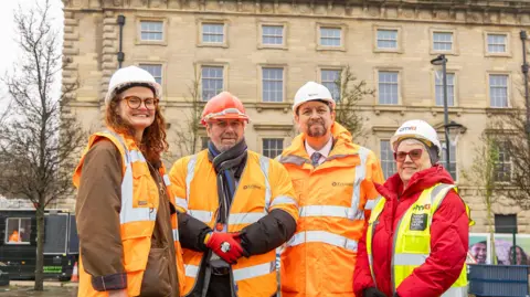 Kirklees Council Four members of Kirklees Council are standing outside in front of the George Hotel in Huddersfield. They are all wearing hard hats and bright orange and yellow safety jackets.