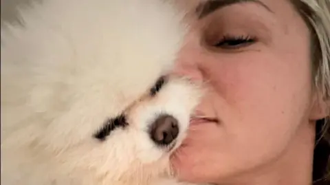 Joanne Wade A close-up of the face of a white pomeranian with its eyes shut, cheek to cheek with Joanne Wade, who has medium-length blonde hair.