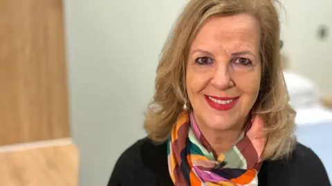 Martin Giles/BBC Caroline Donovan smiling while looking directly at the camera. She has long blonde hair and is wearing a black top and multicoloured scarf.
