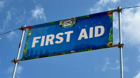 A first aid banner at Kendal Calling. It is blue, with white lettering and has  a Kendal Calling logo. It is set high on poles, against the blue sky. 