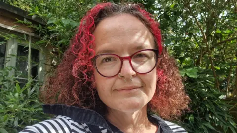 Contributed A woman takes a selfie as she stands outside. She has dark hair with the front sections dyed a vibrant red colour. She has red glasses and a nose ring and is wearing a white and navy striped top. Behind her is a large bush and a window of a building.