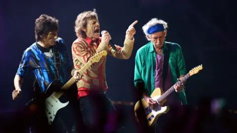 The Rolling Stones are standing on a stage and are wearing colourful clothing. Mick Jagger, who is in the middle, is holding a microphone and is singing, while two people stand either side of him playing guitars.