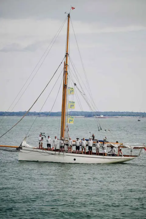 Cowes Week Martin Allen A traditional sailing yacht with crew members standing on deck as it travels across calm water.