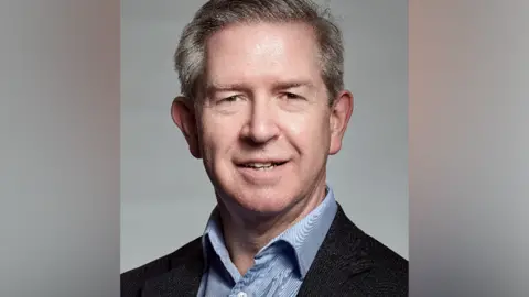 Grainger & Worrall Head and shoulders image of Duncan Eldridge, wearing a blue striped, open-neck shirt under a dark suit jacket