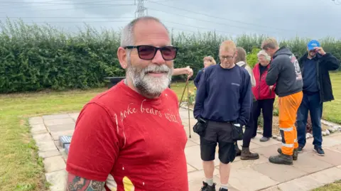 Philip Grimwood, a man with grey hair and a grey beard, is wearing a red T-shirt and sunglasses. He is stood by a memorial. There is a group of people behind him, gathered at the site of a memorial.