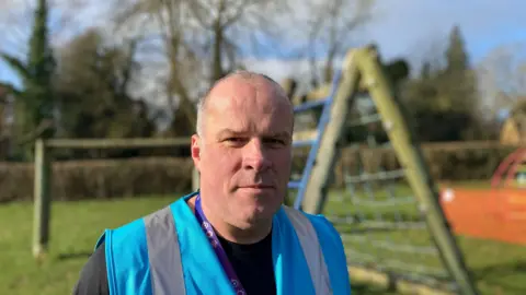 Close up of Clive Sparling in a blue hi-vis jacket. He has grey receding hair. The background  of a play area is slightly blurred. 