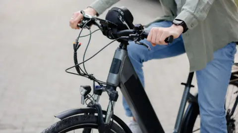 Sandu Herta/Getty Images A person dressed in blue jeans and a green shirt is riding a black e-bike along a well-maintained brick pathway in a park. 