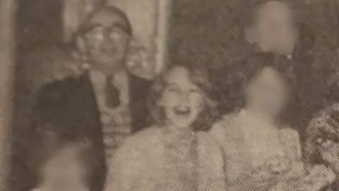 Black and white photograph. On the left is William Barrie who is bald and wearing glasses, tie, jumper and blazer. On the right is Mary with a bob. 