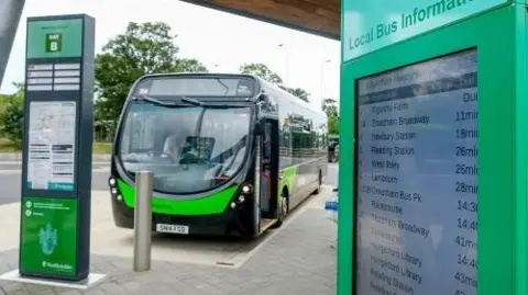 A modern green and black electric-style bus is stopped at a bus station bay next to a tall bus stop sign and a large green information display titled “Local Bus Information.” The bus is parked on a paved platform beneath a wooden roof structure, with trees visible in the background. A route timetable board stands on the left, and the front of the bus is facing the camera. Visible text includes “Bay B” on the bus stop sign and destination information on the timetable display.