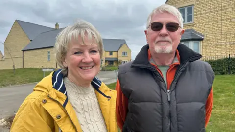 Cath McMurtry (left) and Mike McMurtry (right) looking at the camera smiling while wearing coats, in front of a new housing estate