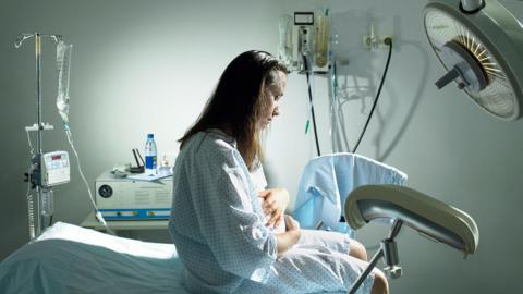 A pregnant woman, sitting on a hospital bed, holds her stomach