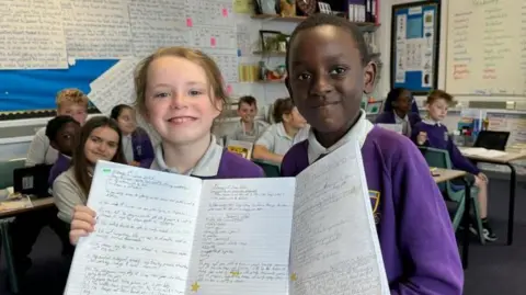Maxwell has blonde hair, tied back, while Rayleigh has short black hair. They are both wearing white polo shirts and purple jerseys, embroidered with the school logo. They are holding their A4-sized workbooks open. Other children are sitting at desks behind them. School work is displayed on the classroom's walls.