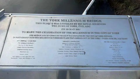 BBC/ Emily Johnson A metal plaque mounted on part of a bridge displays the dedication and contributors to the York Millennium Bridge, with water and greenery visible softly out of focus behind it.
