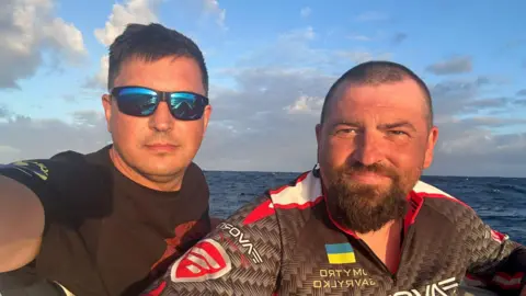 Dmytro Rezvoy, in blue sunglasses and a black T-shirt, and Ivan Havrylko, wearing a shirt with Ukraine's flag on it, in their rowing boat at sea. 