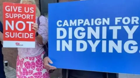 People holding placards, a smaller one on the left reading: "give us support not suicide" and a larger blue one on the right reading: "campaign for dignity in dying".