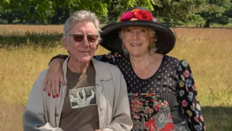 Gavin Petrie and Jan Etherington in a field. He is on the right and wearing sunglasses and a brown top and cream overshirt. Jan has her arm around him and is wearing a big black hat with flowers around the brim 