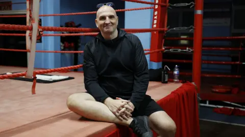 Facundo Arrizabalaga / MyLondon Ray McCallum is wearing a black hoodie, black shorts, black sneakers and glasses. He sits next to a red boxing ring.