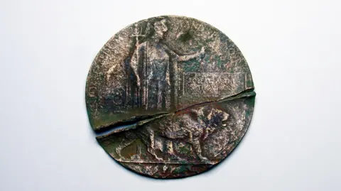 David Stuckey An old bronze plaque. It is circular and split in two. On it is the figure of Britannia, a helmeted female warrior holding a trident and shield, with a lion at her feet. The name of Robert Arthur Thompson is engraved on the plaque within a rectangular border, between Britainnia's outstretched left arm and the lion's head.