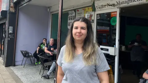 Niya Konakchieva, who is smiling, wearing a grey t-shirt and has long brown hair with blonde highlights, stands outside a cafe where customers can be seen sitting on outdoor chairs. 