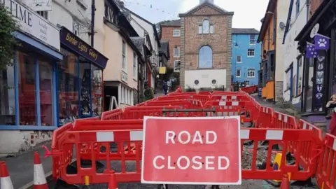 Shropshire Council A red "road closed" sign in front of dozens of red traffic barriers, which have been placed up a street. There are shops either side of the road, and a building at the top of the road with a large rounded window 