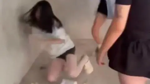 Social media screengrab A blurry screenshot of a girl in a white t-shirt and shorts falling on the ground. Surrounding her are two other girls in black attire.