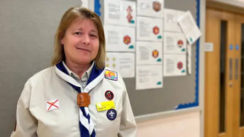 Jo smiles at the camera and is wearing a Scouts uniform. She has short blond hair which comes down to her shoulders. She has several badges on her cream coloured shirt and a neckerchief that is blue and white.