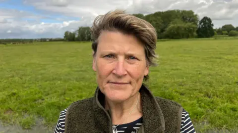 Sarah Dyke, the MP for Glastonbury and Somerset standing in a green field with trees behind. She has sort brown hair and is wearing a brown fleece gilet with a stripey black and white top.