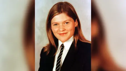 Wilson Family Lorraine Wilson with long brown hair and green eyes as she smiles at the camera wearing her school uniform. She has a black blazer, white shirt and black striped tie on.