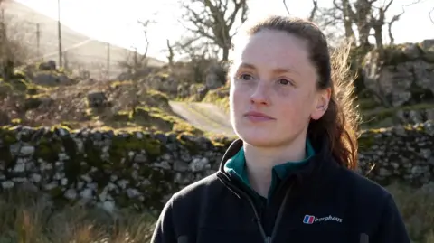 Robyn Wrigley a University of Leeds researcher stands in front of a Yorkshire drystone wall wearing a dark navy zip up fleece. She is white and her brown hair is tied up in a pony tail. The sun is shining in the background highlighting the limestone landscape and vegetation.