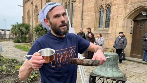 Lee Johnson had brown hair and a brown beard. He is wearing a chef hat and a blue t-shirt which says Tempest on Tithebarn in orange writing. He is holding a frying pan and a silver pan.