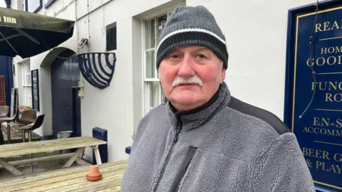 An older man, wearing a grey and black fleece and a grey, black and white beanie hat, stands at the front of a pub, where wooden benches have been put out for patrons