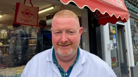 A head and shoulders shot of a Simon Kneafsey standing outside his business M&K Butchers. Mr Kneafsey has short, cropped ginger hair and a very short ginger beard, and wears a white overcoat and a green and blue checked shirt.