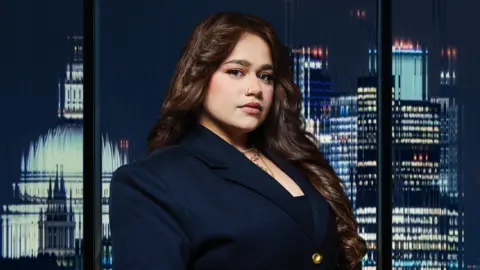 A woman wearing a dark coloured blazer. She is stood in front of a set of glass windows, with a city skyline visible behind her.