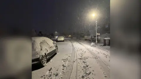 Liv A road covered in white snow. There is a street light on the left and a number of cars covered in snow on the right. 