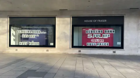 A grey building with two large windows. There is a House of Fraser logo on the right window. Both windows have signs that say "closing down 20% off full price". 