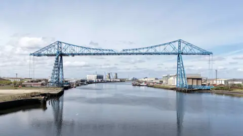 CAV Aerial A view of the Tees Transporter Bridge from further down the river. The large blue structure stretches all the way over the water and is reflected in the surface.
