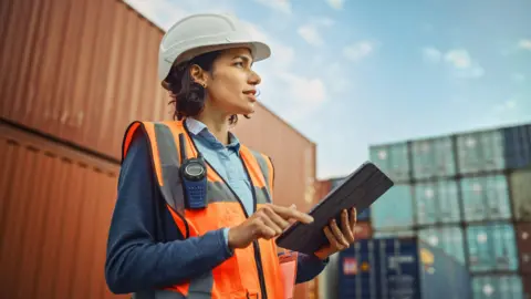 Getty Images A woman with short brown hair wearing orange high vis and a white hard hat holding a tablet while standing in front of shipping containers