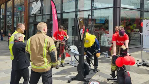 Royal Berkshire Fire and Rescue Service A group of firefighters are working out on exercise machines, while others watch on. In the centre is a large bear-like mascot figure in a firefighter's uniform who is taking part in the exercises. 