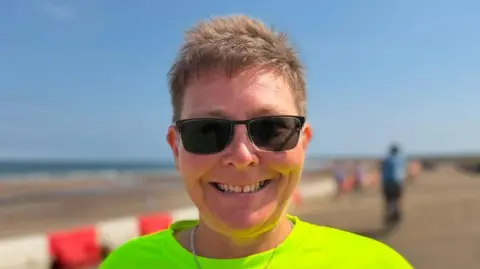 Michele Weedy is wearing dark sunglasses on a beach and smiling. She is wearing a bright green top and has short cropped hair. In the background you can see the beach.