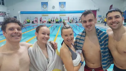 Grantham Rotary Club A group of swimmers stood in front of a pool. Three men can be seen in swimming trunks and two women with towels on. They are all smiling into the camera.