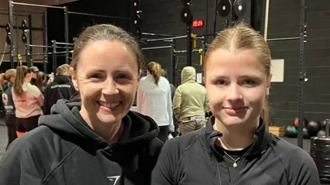 A mother and daugher both with brown hair on ponytails and wearing black gym clothes smile at the camera