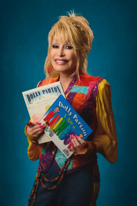 IMAGINATION LIBRARY Dolly Parton in a brightly coloured outfit holding up two of her children's books.