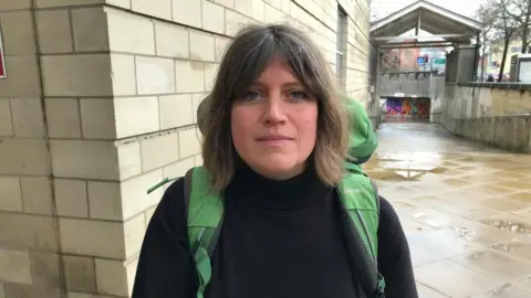 BBC/Sam Read Bethany Mogie with long dark hair, wearing a dark top and carrying a green ruck sack, stands outside a stone-built court building