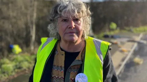 A grey-haired woman with a hi-vis jacket and dark fleece looks at the camera, while litter pickers work at a lay-by in the background