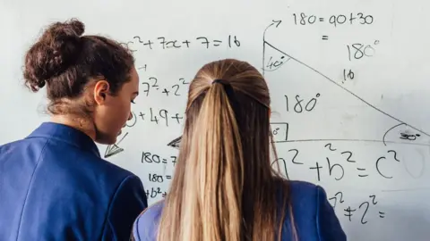 A rear view of two female pupils in school uniform doing a maths problem on a whiteboard