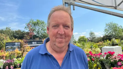 Kevin pictured smiling and looking into the camera. He has grey hair and is wearing a sky blue short sleeved polo shirt. He's stood outside at a garden centre under a white awning, with baskets of bright pink flowers on his right. Behind him are rows of more plants - the sky is bright blue with a few clouds.