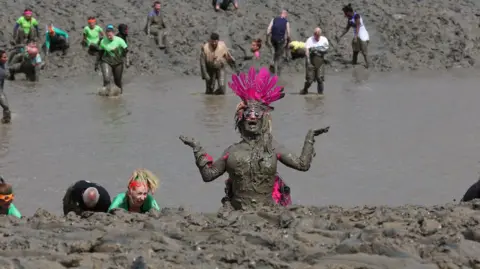 Chris Radburn/Fixedpoint Media A woman with a pink-feathered head-dress is visible over the bank of the estuary. She has her arms bent at the elbows and outstretched at her sides with her palms facing upwards. A stretch of water is visible between the mud in the foreground and the bank in the background. There are about 20 other mud racers visible.