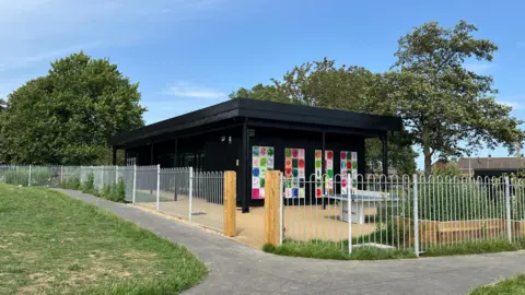 BBC News The purpose-built Cherry Tree Youth Hub. It has silver fencing surrounding it and the building is black with artwork on the sides. Outside you can see a ping-pong table. 