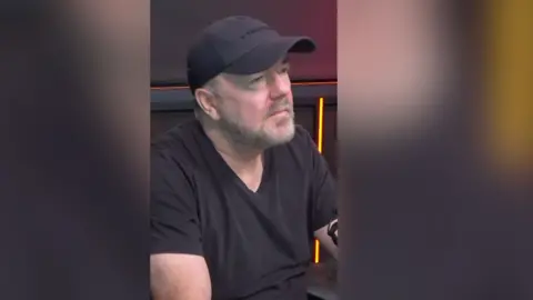 A man in a black cap and black shirt sits.
